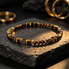 A close-up of a tiger eye bracelet with square gold beads resting on a dark stone surface.
