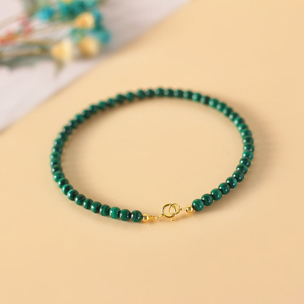 A thin bracelet made of small round green malachite beads with a gold tone clasp on a beige background