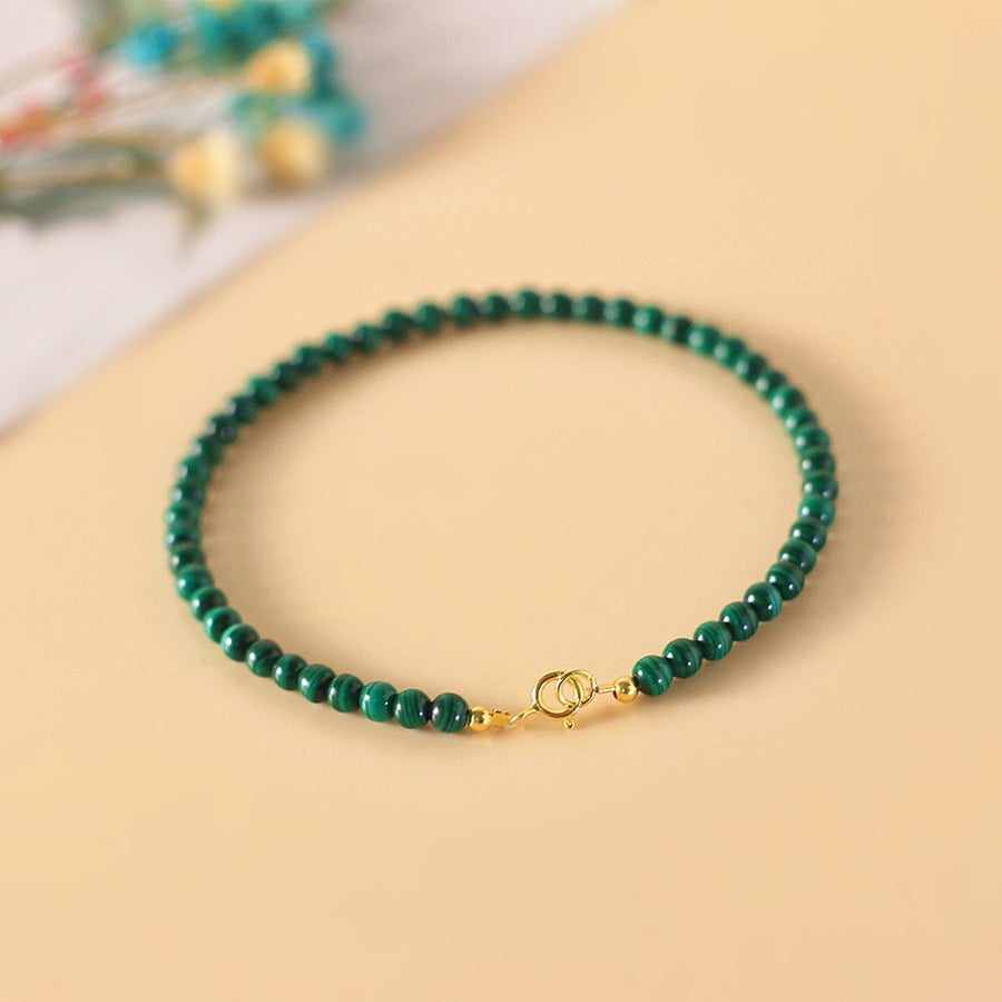 A thin bracelet made of small round green malachite beads with a gold tone clasp on a beige background