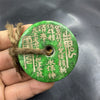 Hand holding a large green jade coin pendant with gold inscriptions and a hemp cord.
