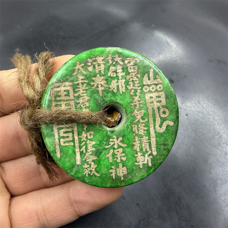 Hand holding a large green jade coin pendant with gold inscriptions and a hemp cord.