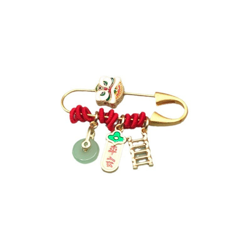 Gold-tone safety pin brooch with lion bead, red cord, and hanging charms including a green circle and ladder.