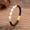 A close up of a black braided leather bracelet with white irregular pearls and gold tone hardware on a wood surface