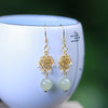 Pair of dangle earrings with gold flower charms and pale green jade beads resting on a white ceramic cup.