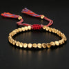 Gold-tone copper bead bracelet with red woven cord and tassels on a black background