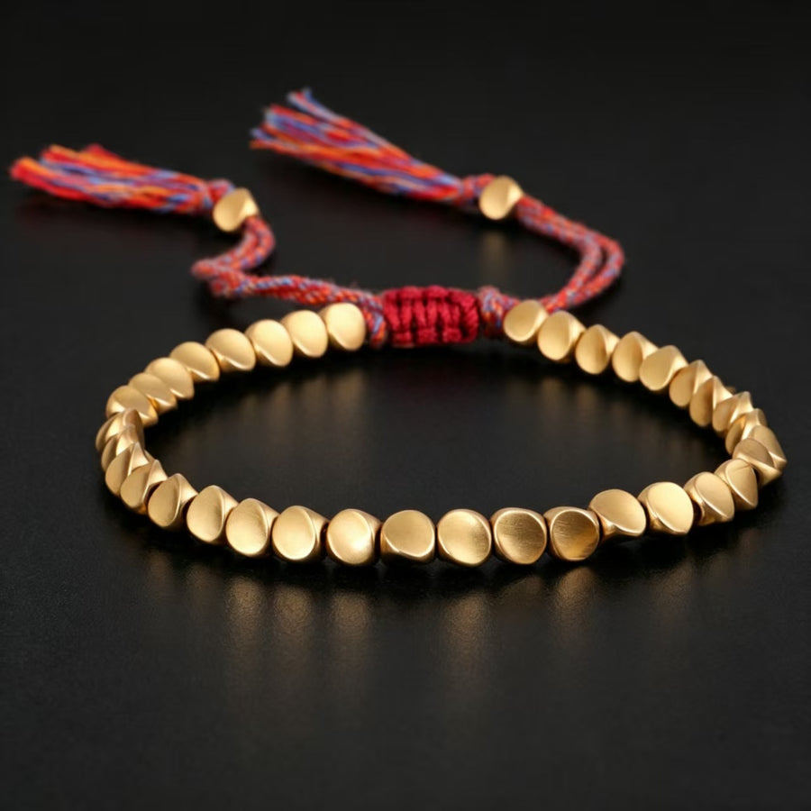Gold-tone copper bead bracelet with red woven cord and tassels on a black background