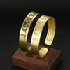 Gold-tone brass open cuff bracelet with engraved Tibetan mantra on a wooden stand