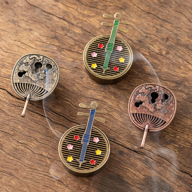 Small round alloy incense burners shaped like a pipa lute and a fan sitting on a wooden surface with smoke rising