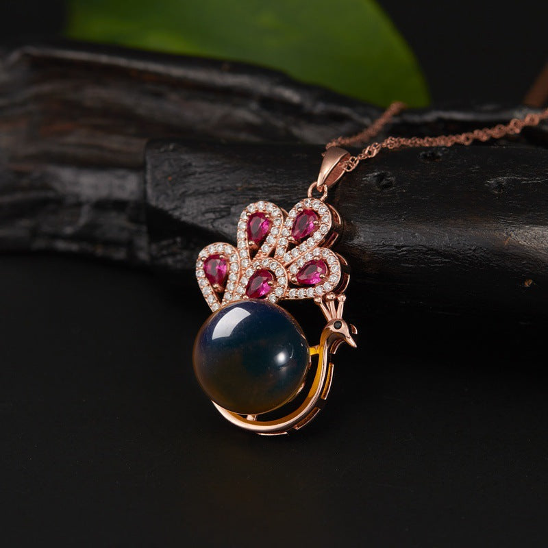 Close-up of a peacock pendant necklace with a blue stone and red gems on a dark surface.