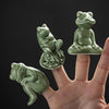 Three small green ceramic frog figurines in different poses held on fingertips