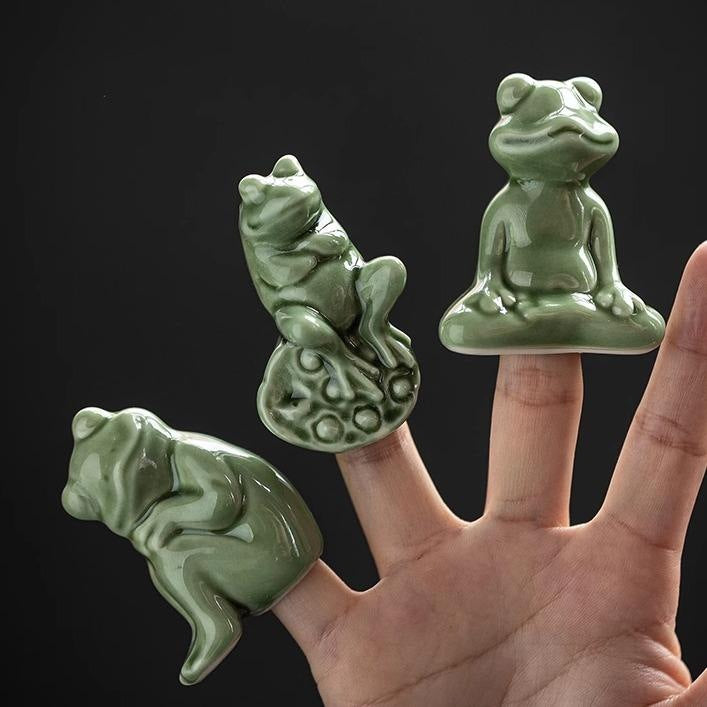Three small green ceramic frog figurines in different poses held on fingertips