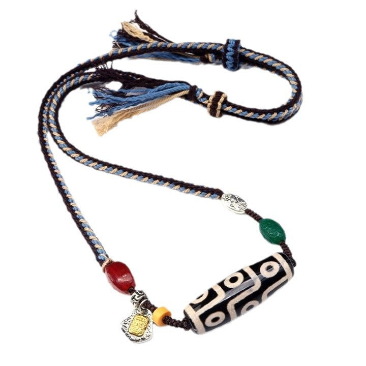 A close-up of a braided cotton rope necklace featuring a black and white Dzi bead pendant with red and green accent stones.