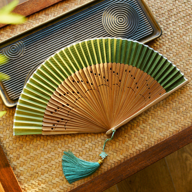 A green and tan Chinese style folding fan with a tassel resting on a woven mat.