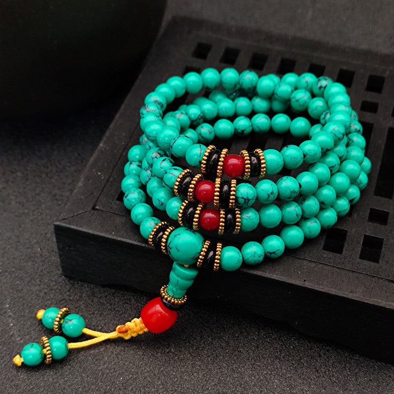 Coiled 108-bead turquoise mala bracelet with red accents and tassel on a dark wooden stand