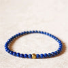 Close up view of a thin blue lapis lazuli beaded bracelet with one gold bead on a textured surface