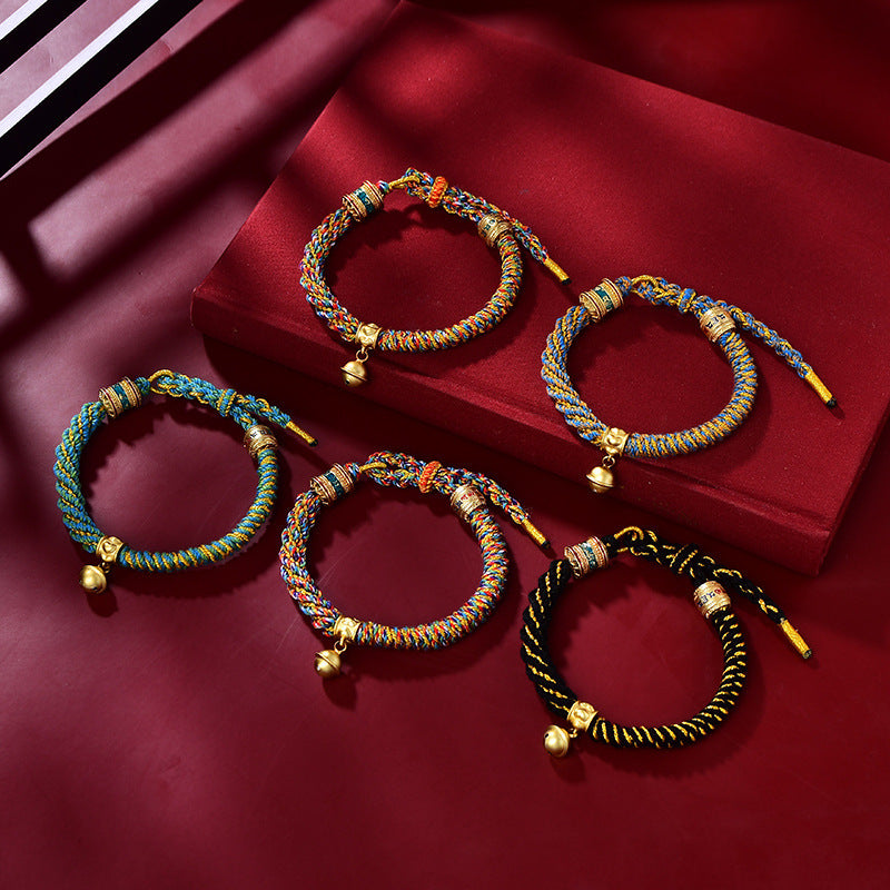 Five colorful handwoven corn knot bracelets with small gold bells arranged on a red surface