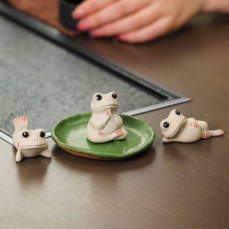 Three small white ceramic frog figurines arranged on a wooden table with a green lotus leaf dish.