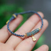 Close up of a blue braided bracelet with light green jade beads and a gold charm held in a hand