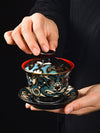Hand holding a black and blue gaiwan teacup with a Peking Opera mask design