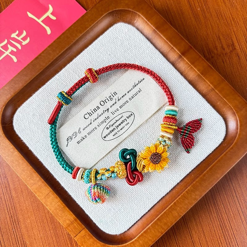 Close-up of a woven red string bracelet with a yellow sunflower charm on a wooden tray