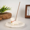 A small white ceramic cat figurine holding an incense stick on a saucer