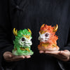 Two hands holding green and orange resin Kirin tea pets