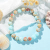 A close-up of a light blue and multi-colored natural stone bracelet resting on a white textured surface with seashells.