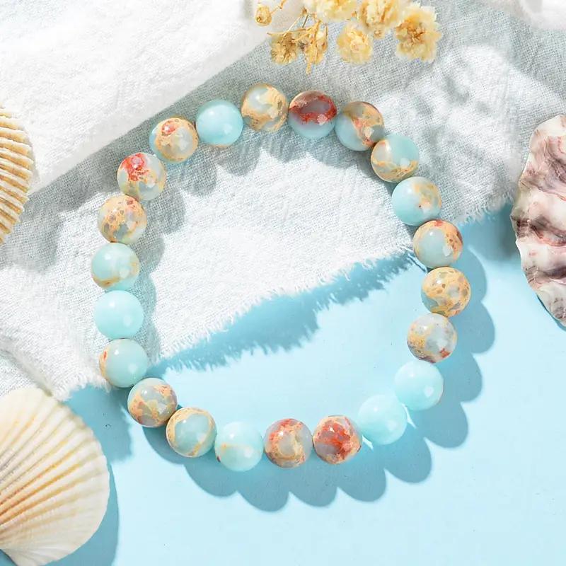 A close-up of a light blue and multi-colored natural stone bracelet resting on a white textured surface with seashells.