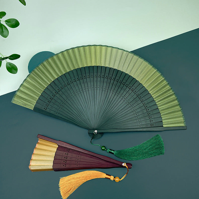 A green and yellow gradient handheld folding fan with a matching green tassel lying on a surface.
