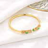 Close up of a gold-tone open cuff bracelet with green stones and filigree detail on a white surface