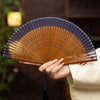 A hand holding an open mini folding fan with hollow carved bamboo ribs and a blue fabric edge.