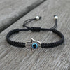 Black woven cord bracelet with a silver tone Hamsa hand and blue evil eye charm on a wooden surface