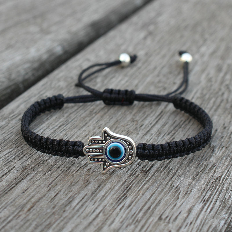 Black woven cord bracelet with a silver tone Hamsa hand and blue evil eye charm on a wooden surface