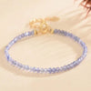 A delicate bracelet made of small faceted purple amethyst beads with a gold-tone clasp on a cream background.