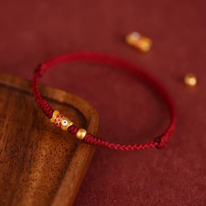 Close up of a braided red string bracelet with a gold koi fish charm on a wooden surface.