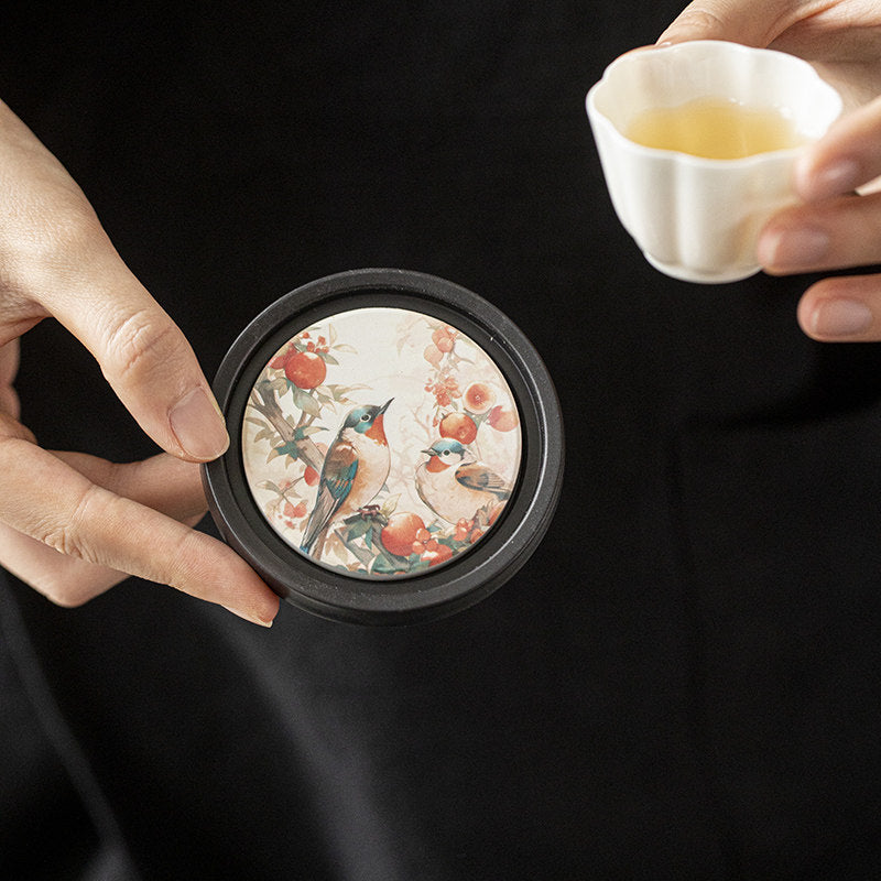 A hand holds a round ceramic coaster with a bird and fruit design near a teacup