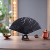 A black bamboo folding hand fan with white ink patterns resting on a wooden stand
