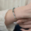 Close-up of a silver bamboo charm on a green and gold braided bracelet worn on a wrist.