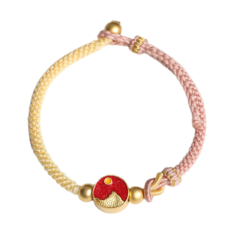 Close-up of a two-tone pink and yellow woven bracelet with gold-tone beads and a red mountain and sea charm.
