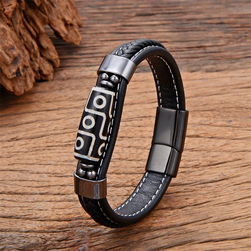 Black braided leather bracelet with a three-eye Dzi bead and silver-tone titanium steel clasp on a wooden surface.