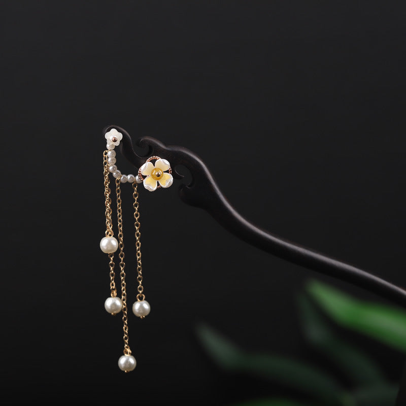 Close-up of a dark wooden hairpin with a white flower and hanging pearl chains on a black background.