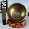 A brass-colored copper singing bowl with mantra engravings resting on a red cushion next to a wooden mallet.