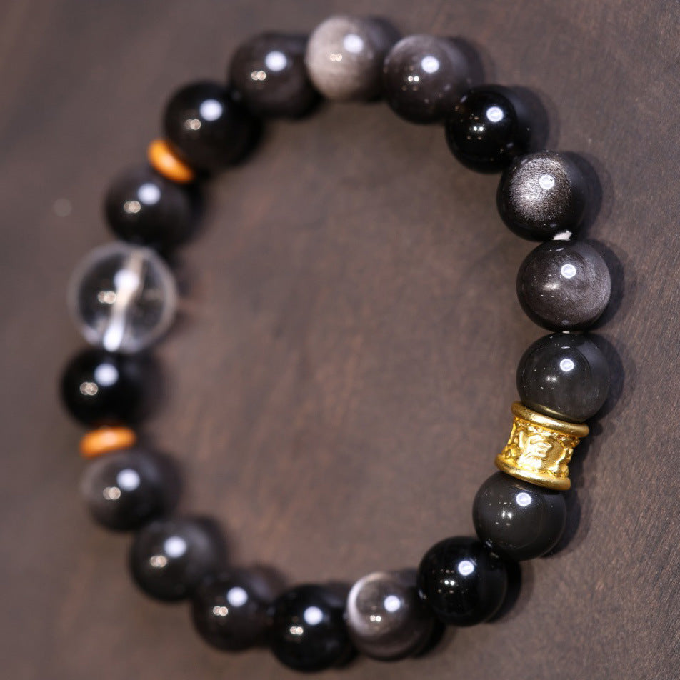 A beaded bracelet made of black silver sheen obsidian with a gold-tone charm resting on a wooden surface