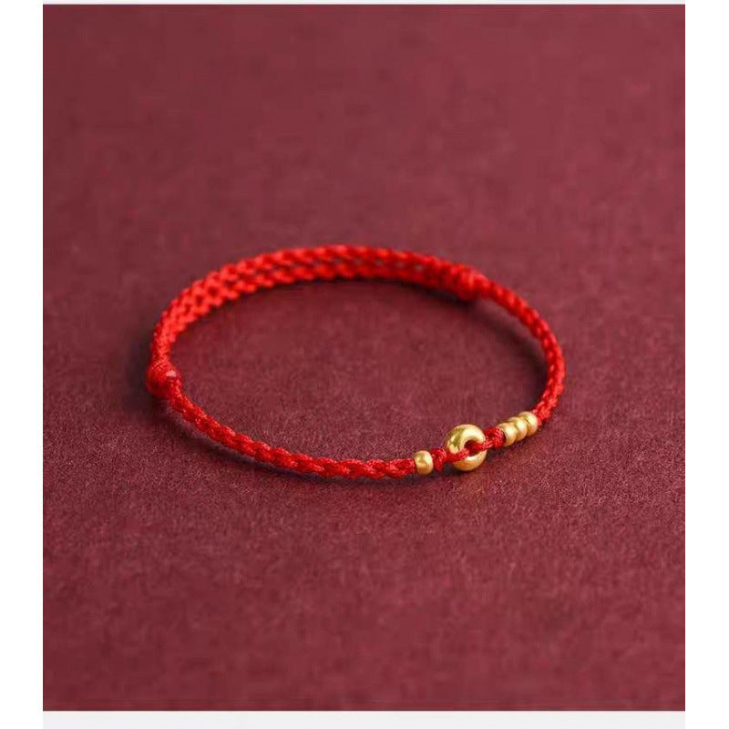 A braided red cord bracelet with a small gold-tone ring charm on a dark red background