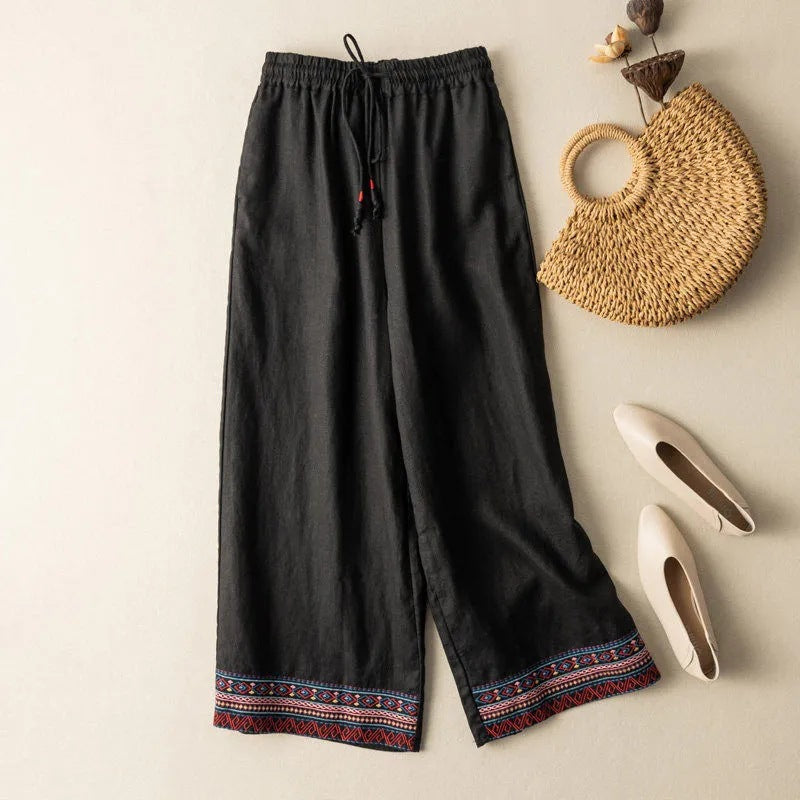 Black wide leg linen pants with colorful embroidery at the hem laid flat on a beige surface