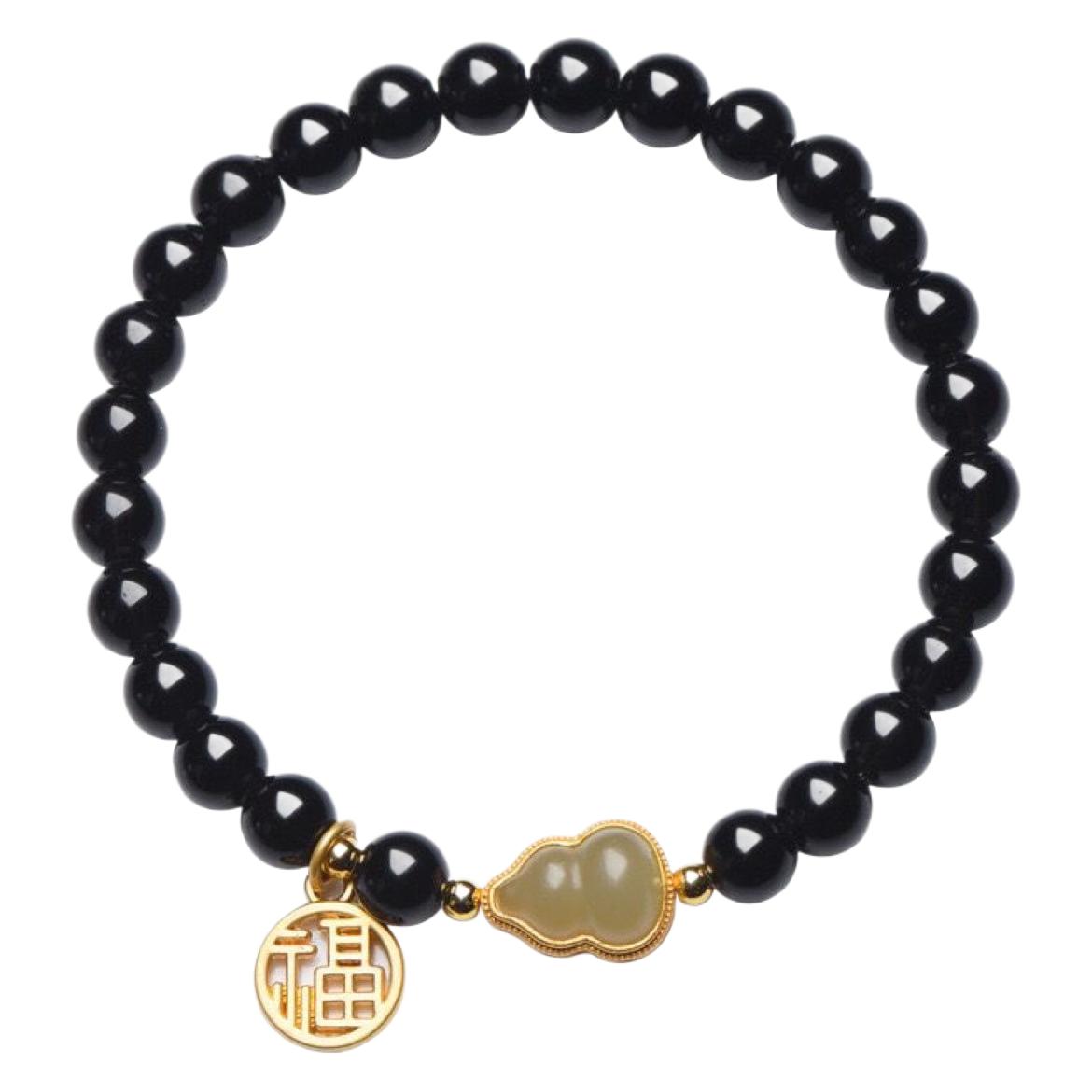 Close up of a bracelet with black beads, a jade gourd charm and a gold double happiness pendant on a wrist