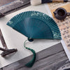 Green woven fabric folding fan with bamboo ribs and tassel on a wooden table