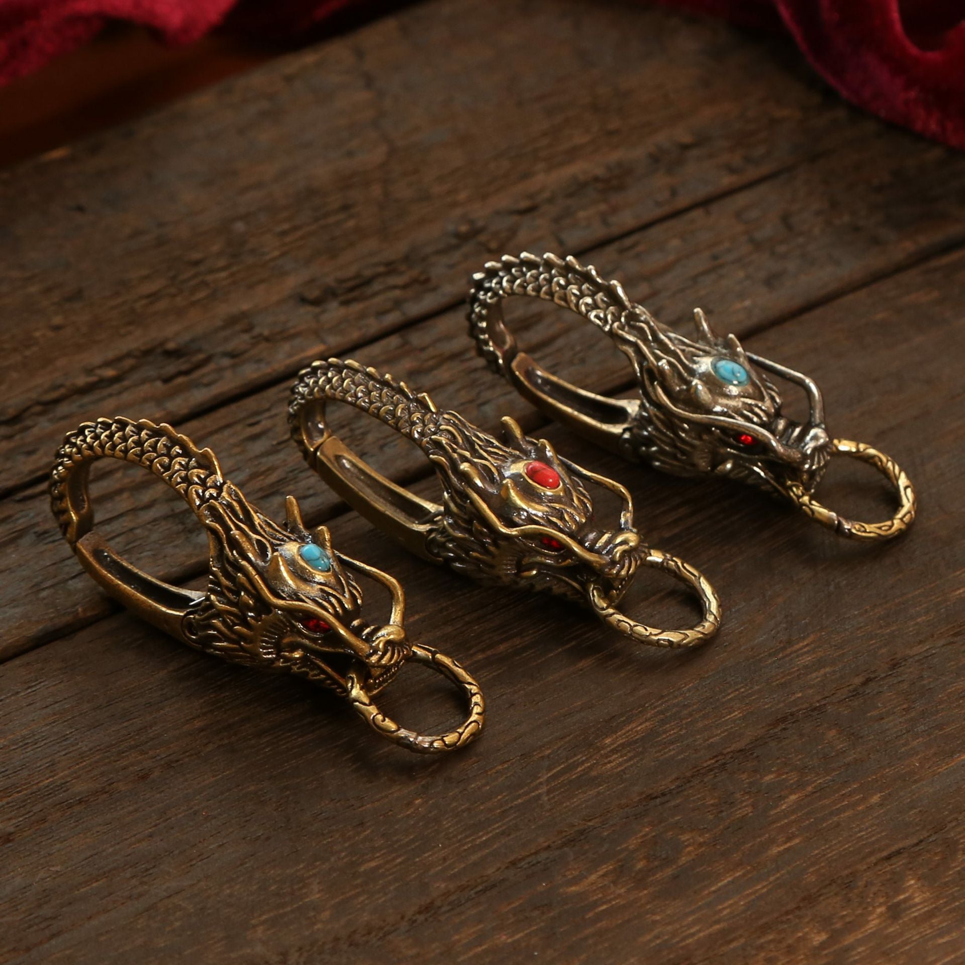 Three brass dragon head keychains with red and blue stones on a wooden surface