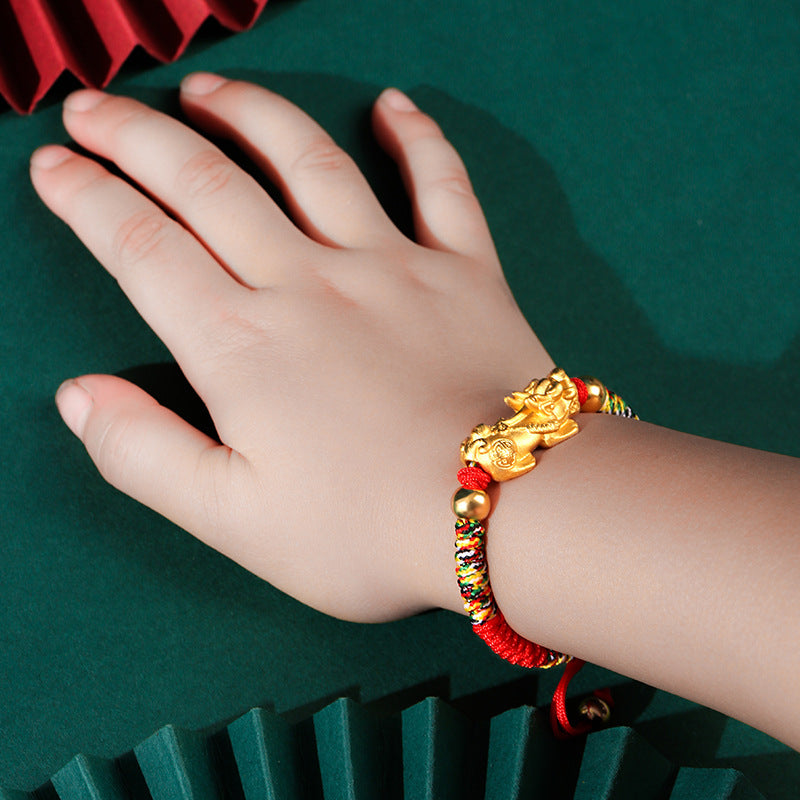 Child's hand wearing a red braided bracelet with a gold-tone Pixiu charm