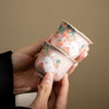 Two hands holding small pink ceramic cups with painted white rabbits and strawberries